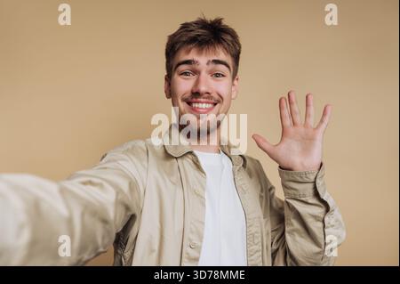Ein junger Mann lächelt und winkt mit der Hand, während er ein Selfie macht. Er trägt ein helles Hemd und hat kurze Haare, steht vor einem einfachen Beige Stockfoto