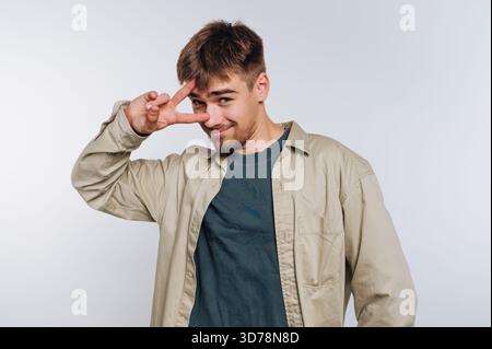 Ein junger Mann posiert spielerisch mit einem Friedenszeichen in lässigem Outfit. Sein Ausdruck zeigt Freundlichkeit und Zugänglichkeit gegenüber einem einfachen Stockfoto