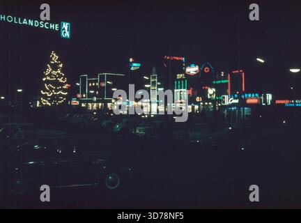 Utrecht, Niederlande, 1964 – nächtliche Stadtlandschaft mit hell beleuchteten Neonwerbeschildern, Weihnachtsdekorationen und beleuchteten Geschäften. Ein großer, geschmückter Baum steht in der Nähe geparkter Autos, während die leuchtenden Umrisse von Kinos, Cafés und Ladenfronten den Platz erleuchten. Das Foto fängt die lebendige Atmosphäre des Nachtlebens von Utrecht in den 1960er Jahren ein, gefüllt mit Farben, Schildern und festlichen Winterlichtern. Stockfoto