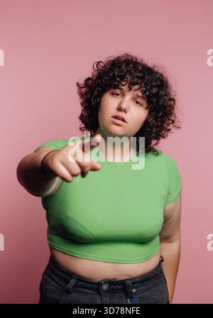 Ein junger Mensch mit lockigen Haaren steht selbstbewusst, trägt ein grünes T-Shirt und eine schwarze Hose. Sie zeigen auf den Betrachter und erzeugen ein kühnes und Stockfoto