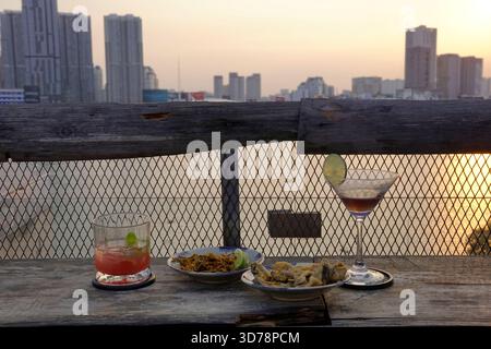 Bunte Cocktails und Snacks auf einem Holztisch mit Blick auf den Fluss und die Skyscapper der Stadt Stockfoto