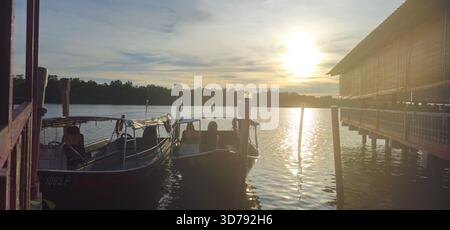 Golden Hour Blick auf zwei traditionelle Boote, die an einem Holzsteg anlegen, mit einem Stelzengebäude und üppigem Waldhintergrund bei Sonnenuntergang oder Sonnenaufgang. Stockfoto