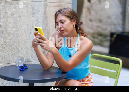 Eine junge Frau, die draußen an einem Café-Tisch sitzt und auf ihrem Smartphone surft Stockfoto