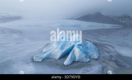 Aus der Vogelperspektive auf einen markanten blauen Eisberg, der in der Gletscherlagune liegt, einen ruhigen Tanz aus Eis und Wasser unter einem nebeligen Himmel, Jokulsarlon, Sveitarfelagid Ho Stockfoto