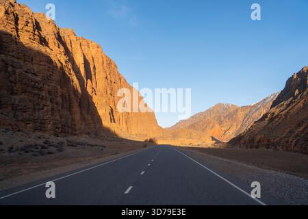Eine leere, gerade Asphaltstraße durchquert ein schmales Tal, flankiert von hohen orange-roten Felsklippen in einer tiefen Bergschlucht Stockfoto