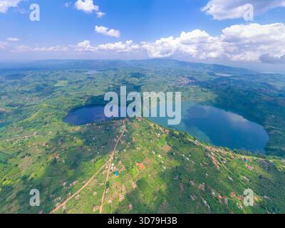 Blick aus der Vogelperspektive auf die Twin Crater Lakes Kyema und Kamweru, eingebettet in einen Wandteppich aus grünen Hügeln unter einem blauen Himmel, im Kontrast zum dunklen und hellen Wasser, Stockfoto