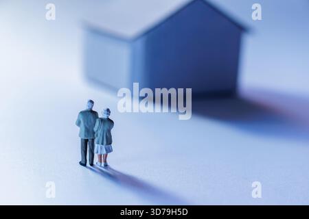 Ältere Paarfiguren stehen zusammen in der Nähe eines Miniaturhauses, während sie ihre Erinnerungen in einer weichen, traumhaften Umgebung reflektieren Stockfoto