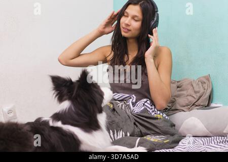 Eine junge Frau genießt Musik mit Kopfhörern, lächelt und macht eine Geste mit einer Hand, während der Border Collie auf dem Boden sitzt und sie beobachtet. Stockfoto