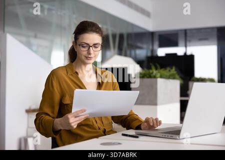 Positive junge Buchhalterin in Brille bei der Prüfung von Firmenfinanzunterlagen Stockfoto