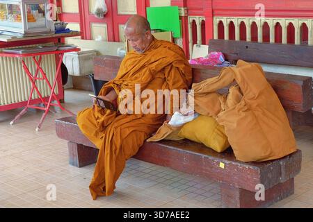 Buddhistischer Mönch für ältere Reisende, der traditionelle orangene Zeremonie trägt und sperriges Gepäck auf der Bank sitzt und auf seinen wartenden Zug blickt Stockfoto