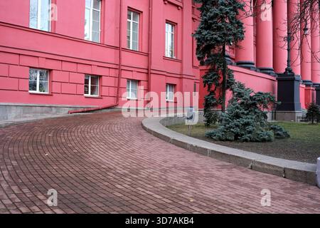 Ein gewundener Backsteinpfad führt durch einen ruhigen Garten neben leuchtend rosa Wänden. 24. November 2025 Kiew, Ukraine, Schewtschenko-Universität Stockfoto