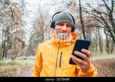 Ein lächelnder Mann mittleren Alters genießt im herbstlichen Jogging-Stadtpark Musik über Kopfhörer, während er im Audio-Streaming-Service Playlisten durchsucht Stockfoto