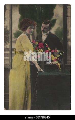 Eine Frau in gelbem Kleid steht an einem offenen Fenster neben einem Tisch mit Blumen, während ein Mann im Anzug sie von hinten ansieht. Stockfoto