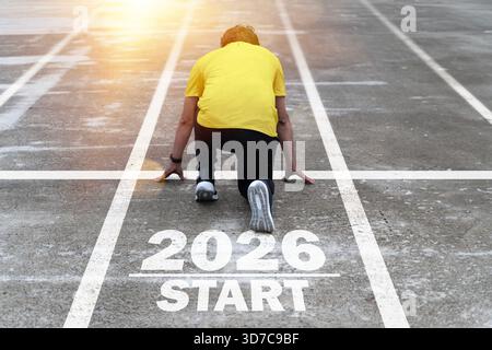 Ein Mann kniet in einer Startposition auf einer Laufstrecke, die mit 2026 Starttext markiert ist, was Neuanfänge, Entschlossenheit, Fortschritt, Ehrgeiz symbolisiert. fut Stockfoto