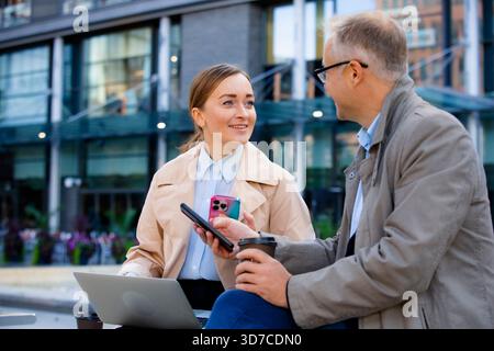 Fachleute halten Geschäftstreffen im Freien mit Laptop und Telefon in städtischer Umgebung ab. Vernetzung und Diskussion. Stockfoto
