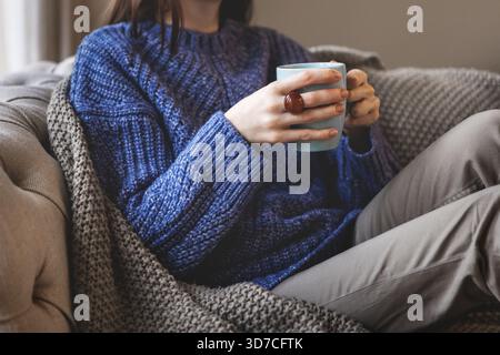 Weibliche Hände halten blauen Becher, während sie in einem übergroßen Strickpullover und einer warmen Decke sitzen. Gemütliche Herbst- oder Winteratmosphäre im Innenbereich mit Komfort Stockfoto