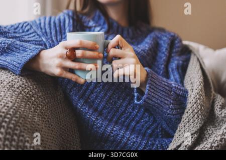 Frau, die sich auf dem Sofa entspannt und eine warme Tasse Kaffee oder Tee trinkt. Gemütlicher Komfort zu Hause, Hygge Lifestyle. Freizeit, Erholung, Wochenende im Winter oder Herbst Stockfoto