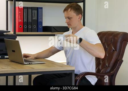 Ein entspannter Geschäftsmann tippt auf einem Laptop und trinkt Kaffee im modernen Arbeitsbereich mit Regalen und Dokumenten. (92 Zeichen) Stockfoto