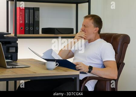 Ein Mann mittleren Alters in weißem T-Shirt sitzt am Holzschreibtisch und denkt über Papiere in blauem Ordner mit Laptop und Kaffeetasse in der Nähe nach. Stockfoto