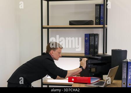 Ein fokussierter Mann beugt sich vor, um einen schwarzen Multifunktionsdrucker zu inspizieren, der auf einem Holzregal in einer modernen Büroumgebung mit Ordnern und einem Laptop platziert ist Stockfoto