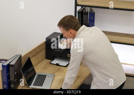 Junge Profi im Hemd beugt sich am Holzschreibtisch nach vorne und trinkt aus einem weißen Becher neben dem ausgeschalteten Laptop und dem schwarzen Drucker in einem hellen Bürobereich. Stockfoto