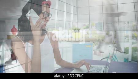 Sprechende Frau in weißem Oberteil, Brille, rotem Stirnband, das Telefon hält und den Laptop im Büro erreicht Stockfoto