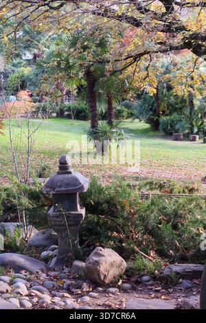 Ruhige japanische Gartenszene im Herbst mit einer traditionellen Steinlaterne umgeben von Palmen, grünem Gras, Felsen und bunten gefallenen Blättern. Stockfoto