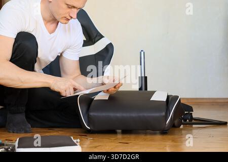 Ein fokussierter Mann in weißem T-Shirt kniet neben dem Sitz des schwarzen Lederspielstuhls und liest sorgfältig die Montageanleitung auf dem Boden. Stockfoto
