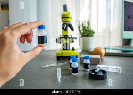 Eine Person, die ein kleines Probenröhrchen vor einem gelben Mikroskop hält, mit mehr wissenschaftlichen Vorräten auf dem Tisch. Das Konzept der naturwissenschaftlichen Bildung und Stockfoto