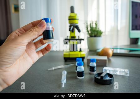 Eine Person, die ein kleines Probenröhrchen vor einem gelben Mikroskop hält, mit mehr wissenschaftlichen Vorräten auf dem Tisch. Das Konzept der naturwissenschaftlichen Bildung und Stockfoto