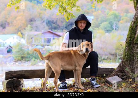 Ein junger Mann in einem schwarzen Hoodie streichelt sanft einen braunen Hund neben sich auf einem Holzstamm am Fluss, umgeben von lebhaften Herbstbäumen und Laub in einer Erbse Stockfoto