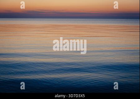 Wunderschöne Meereslandschaft in der Dämmerung mit glatter Wasseroberfläche, pastelloranger lila Himmelsreflektion, ruhiger Horizont nach Sonnenuntergang. Friedlicher, abstrakter natürlicher Hintergrund Stockfoto