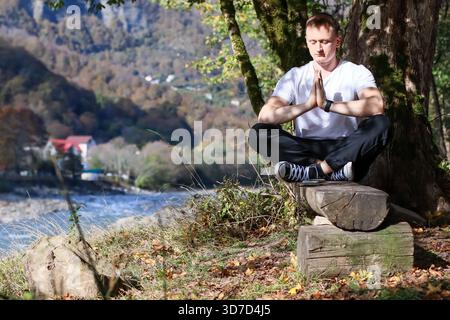 Junger Mann sitzt auf Holzholz unter Baum mit überkreuzten Beinen, Hände in Namaste Position auf Gesichtshöhe, Augen gerichtet nach vorne, malerischer Hintergrund des Flusses, Hügel Stockfoto