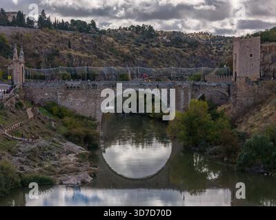 Aus der Vogelperspektive der alten Alcantara-Brücke erhebt sich anmutig über den ruhigen Fluss Tejo unter einem bewölkten Himmel, Toledo, Kastilien-La Mancha, Spanien. Stockfoto
