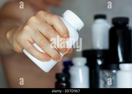 Männliche Hand, die aus einer weißen Supplementflasche mit verschwommenen Nährstoffgläsern dahinter gießt. Athletic man gießt Kapseln oder Pulver aus der weißen Pillenflasche mit Stockfoto