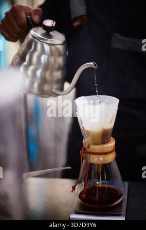 Bali, Indonesien, 18. November 2015. Nahaufnahme eines Mannes, der heißes Wasser aus dem Schwanenhals-Wasserkocher in den Papierfilter mit gemahlenem Kaffee in einer Glaskaffeemaschine gießt und einen frischen, übergießenden Kaffee zubereitet Stockfoto