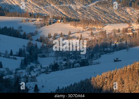 Aus der Vogelperspektive auf ein schneebedecktes Tal mit verstreuten Häusern und goldenem Sonnenlicht, das die Hügel küsst, Chocske vrchy, Prosiek, Zilinsky kraj, Slowakei. Stockfoto
