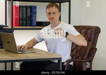 Junger Mann in weißem T-Shirt, der einen weißen Becher hält, während er am Laptop am Holztisch im Büro arbeitet Stockfoto