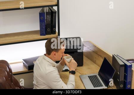 Der Profi nimmt einen Schluck von einem weißen Becher am Schreibtisch mit Drucker, offenem Laptop, Dokumentenakten und Regalen in hellem Innenbereich. Stockfoto