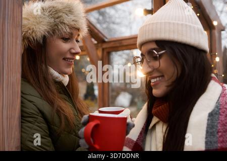 Zwei Frauen genießen sich Gesellschaft, gewickelt in gemütliche Outfits, während sie draußen warme Getränke trinken. Stockfoto