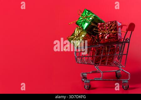 Ein Einkaufswagen voller Geschenkkartons auf rotem Hintergrund. Weihnachtseinkäufe, Weihnachtsverkäufe und Weihnachtskäufe. Stockfoto