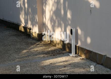 Kompaktes weißes Elektroauto-Ladegerät an einer hellen Wand mit gefliestem Sockel, das Schatten auf Betonpflaster im Freien wirft. Stockfoto