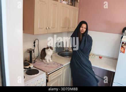 Eine indische Frau umarmt sich in einer dunklen Decke, steht in einer kleinen, einfachen Küche neben einer großen Katze, die auf der Theke sitzt. Einfache Schränke, elektrisch Stockfoto