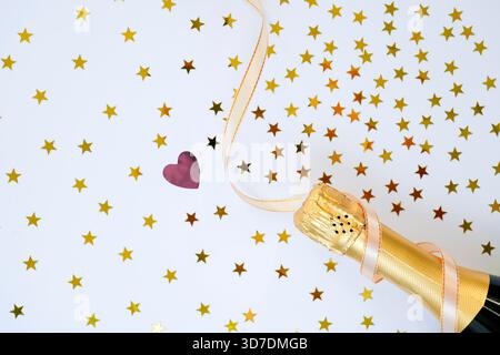 Eine Flasche Sekt ist mit einem rosafarbenen Band gebunden, umgeben von goldenen Sternen und einem roten Herzsymbol, die eine fröhliche Stimmung für ein festliches G-Stück schaffen Stockfoto