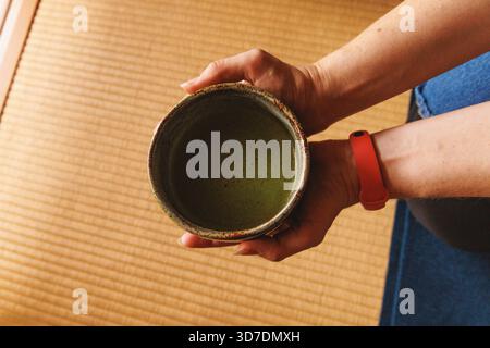 Die Hände halten eine Keramikschale mit aufschäumendem Matcha-Tee über einer Tatami-Matte. Die Szene deutet auf einen ruhigen Moment während eines japanischen Teerituals hin. Stockfoto
