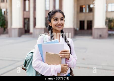 Der Student lächelt, während er Bücher hält und an einem sonnigen Tag vor einem Universitätsgebäude steht Stockfoto