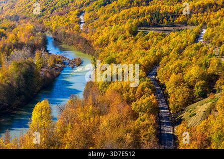 Aus der Vogelperspektive des Venezikos River, der sich durch den lebhaften Herbstwald in Westmakedonien, Griechenland, mit einer parallelen Straße, die in das natürliche Land mündet Stockfoto