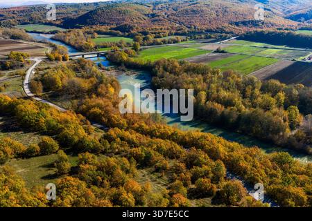 Aus der Vogelperspektive des Venezikos River, der sich durch den lebhaften Herbstwald in Westmakedonien, Griechenland, mit einer parallelen Straße, die in das natürliche Land mündet Stockfoto