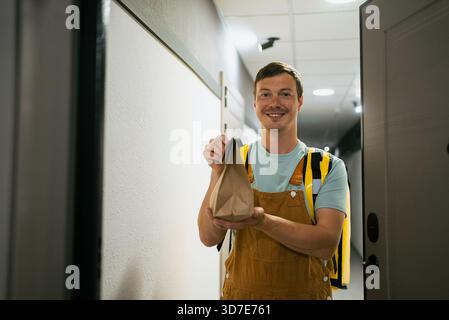Lächelnder Liefermann mit gelbem isoliertem Rucksack, der Fastfood-Mahlzeiten in einer Papiertüte in eine Wohnung bringt und einen kontaktlosen Hausversand vor der Tür des Kunden bietet Stockfoto