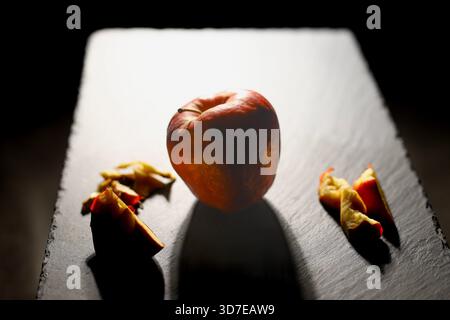 Ganzer Apfel von hinten dramatisch beleuchtet, umgeben von abgeschnittenen Schälstücken auf einem Schieferbrett. Minimalistisches Stillleben betont Textur und Natürlichkeit Stockfoto